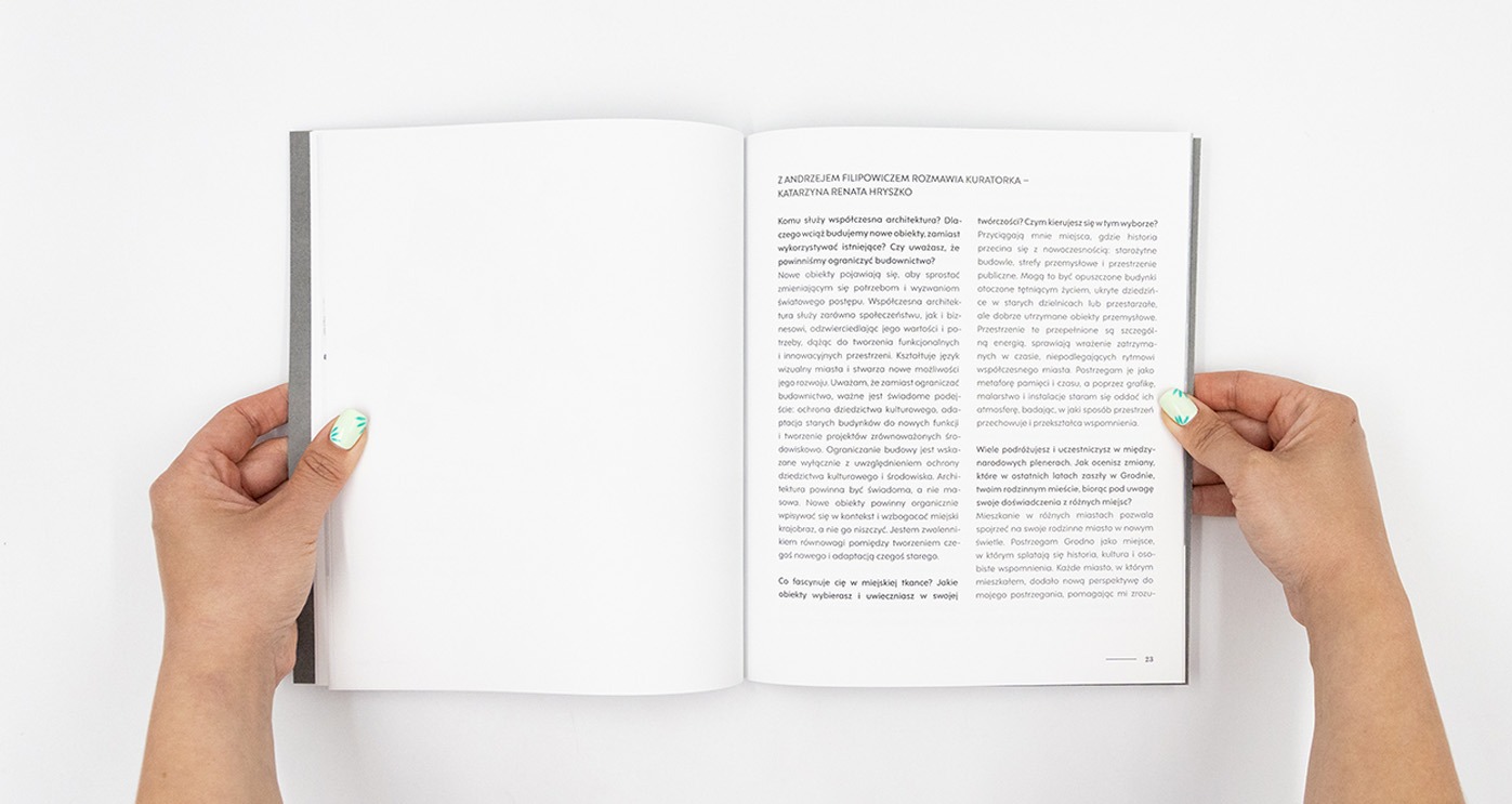Otwarta książka z tekstem o architekturze, trzymana przez osobę z pomalowanymi paznokciami. Białe tło, stonowana kolorystyka. Układ tekstu czytelny, minimalistyczny design.