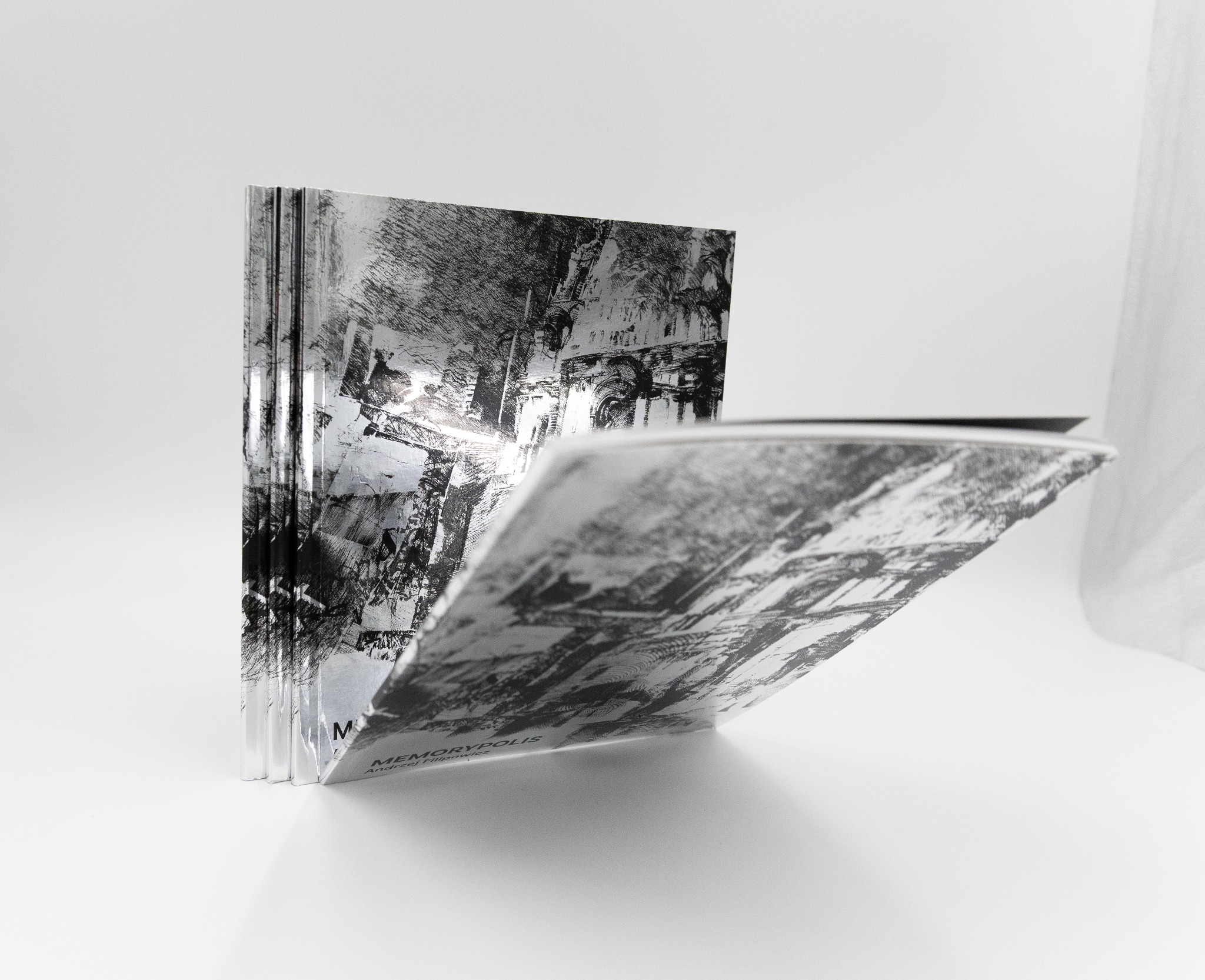 Otwarta książka 'Memorypolis' z abstrakcyjną grafiką miejską w odcieniach szarości, ustawiona na białym tle. Widoczny grzbiet i fragment okładki.