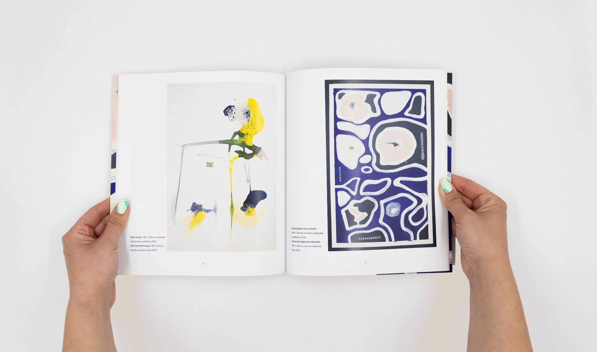 Otwarta książka z abstrakcyjnymi obrazami w kolorach żółtym, niebieskim i fioletowym, trzymana przez dłonie z zielonym manicure na białym tle.