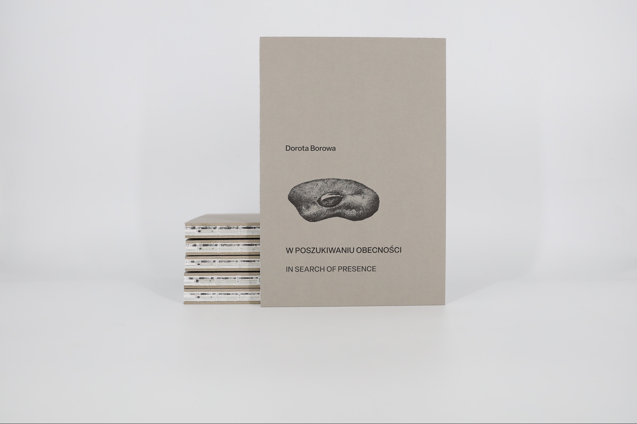 Katalog wystawy Doroty Borowej: szara okładka z grafiką, tytuł 'W poszukiwaniu obecności', obok stos katalogów na białym tle. Projekt Ewa Pietruszko.