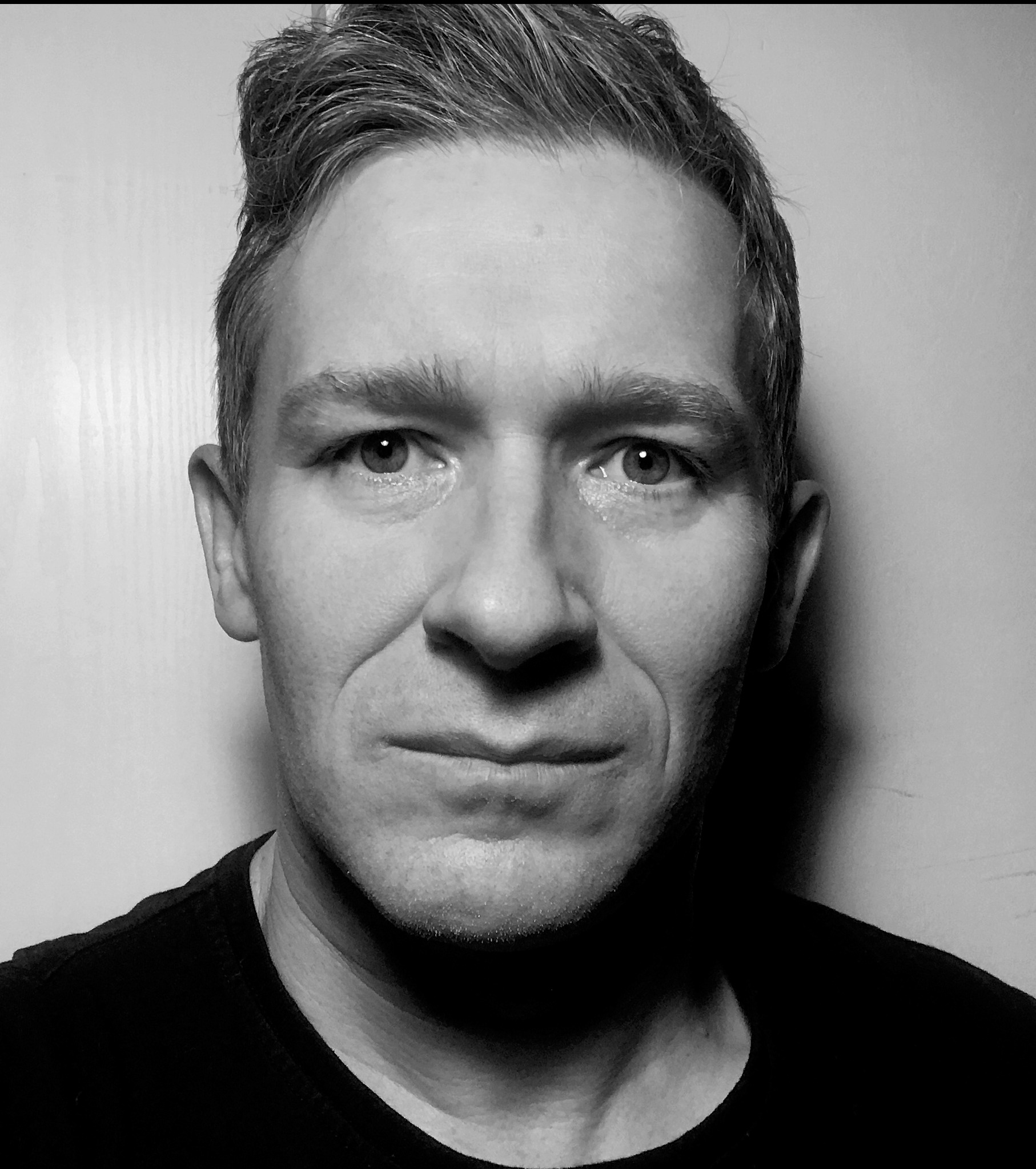 Czarno-biały portret mężczyzny z krótkimi włosami, skierowanego prosto w obiektyw, z delikatnym uśmiechem i widocznymi zmarszczkami wokół oczu. Neutralne tło.