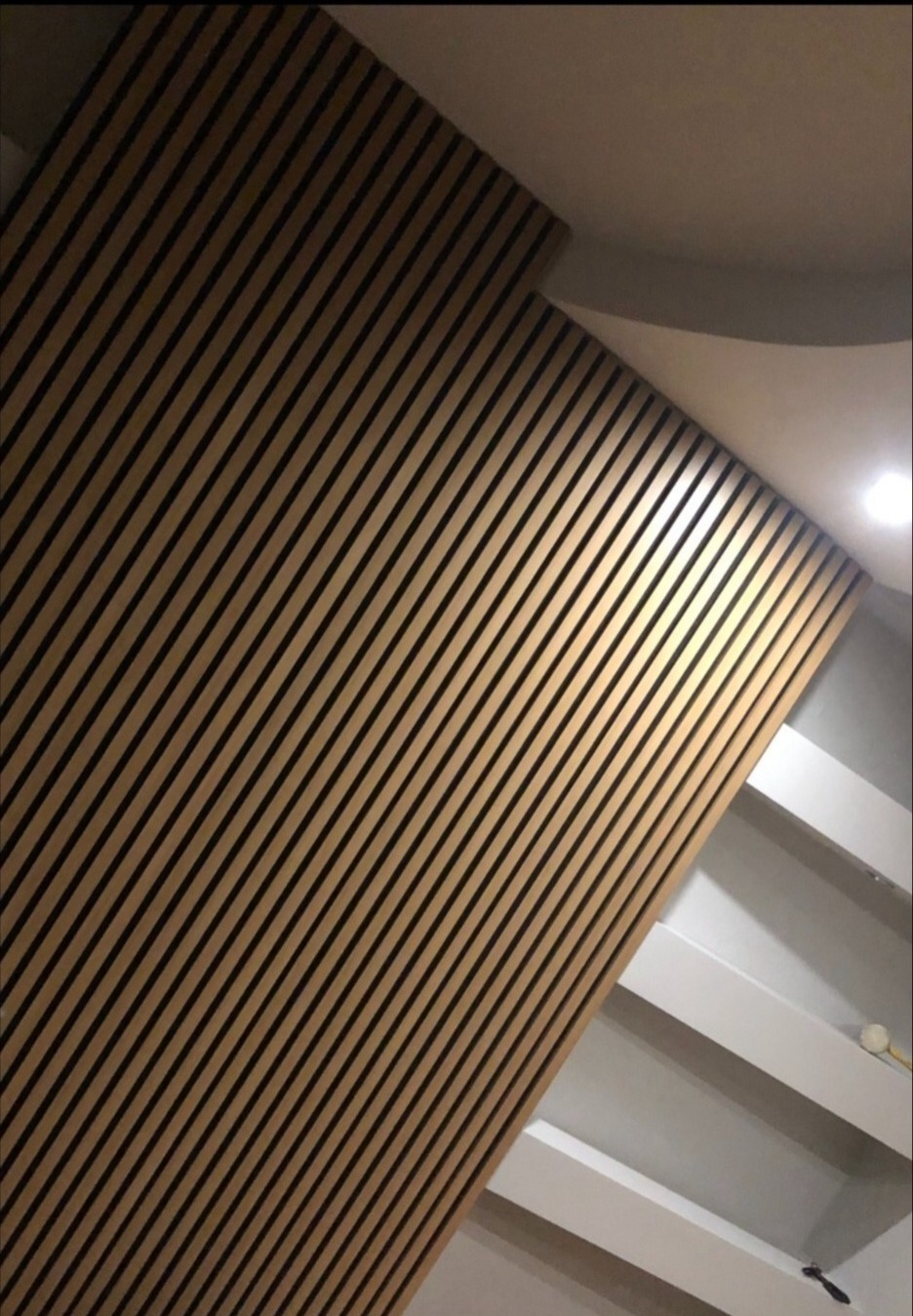 Drewniana ściana lamelowa pod skosem, z widocznymi czarnymi paskami, przylegająca do białych schodów i sufitu. Nowoczesny design w wykończeniu wnętrza.