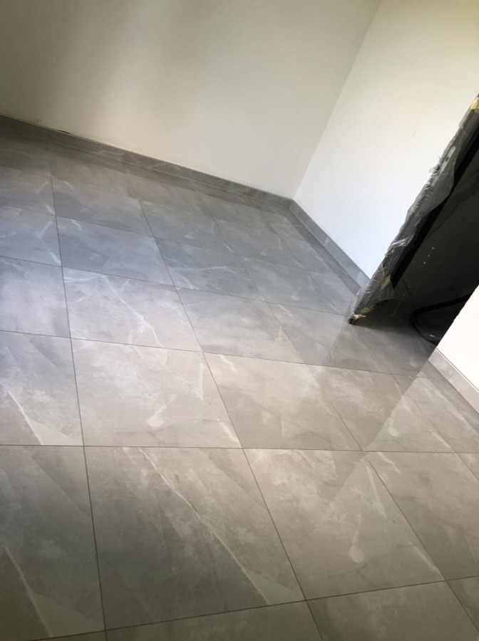 Ukończona podłoga z szarych płytek imitujących marmur, ułożonych w regularny wzór w rogu pomieszczenia z białymi ścianami. Widoczne fugi.
