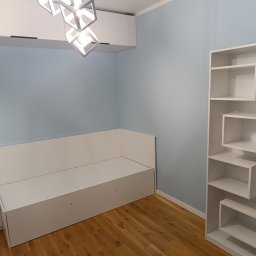 NOWY WYMIAR- Studio Projektów Justyna Matulewicz - Nowoczesne wnętrze pokoju z zabudową meblową: biała szafa, narożna kanapa w zabudowie i regał o geometrycznym designie na drewnianej podłodze. Błękitna ściana.