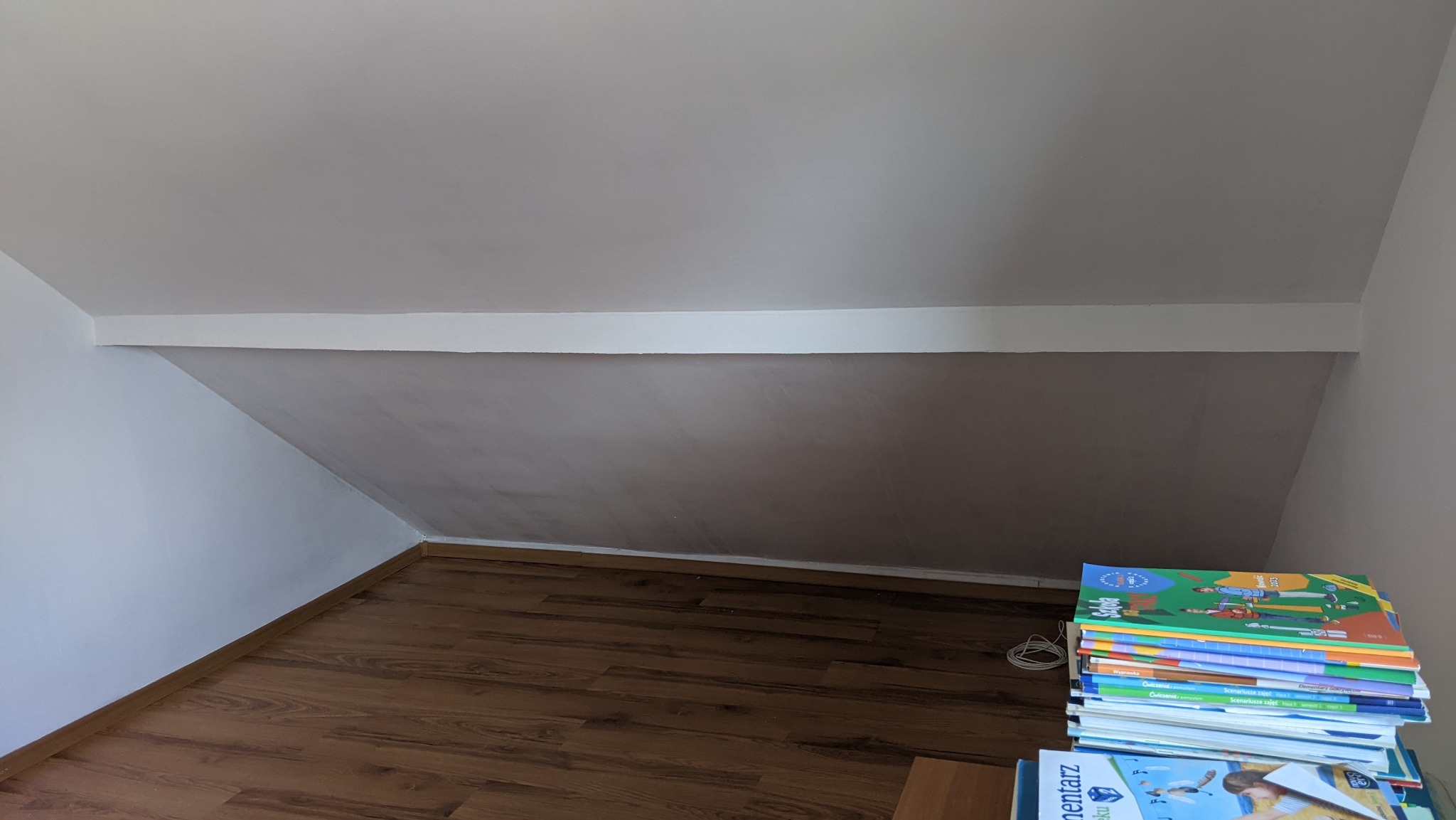 Poddasze po remoncie: świeżo pomalowane ściany w odcieniach bieli i szarości, drewniana podłoga. Stos książek w rogu dodaje wnętrzu przytulnego charakteru.