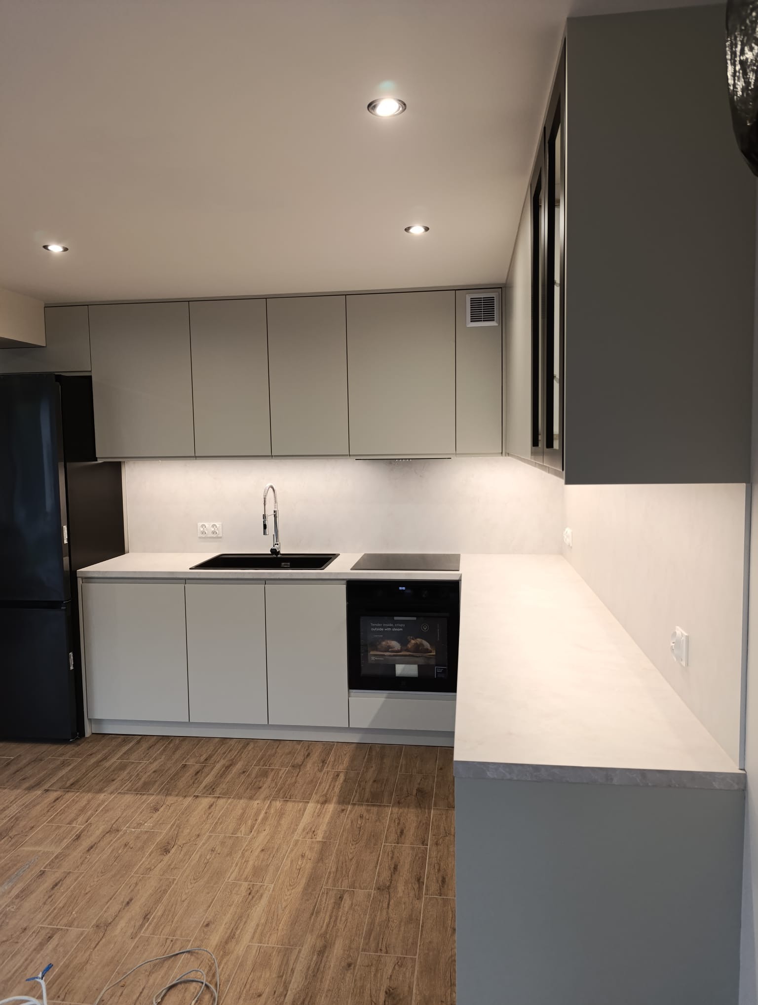Nowoczesna kuchnia w odcieniach szarości z drewnopodobną podłogą. Minimalistyczne szafki, czarny zlew i sprzęt AGD. Subtelne oświetlenie LED podkreśla blat.