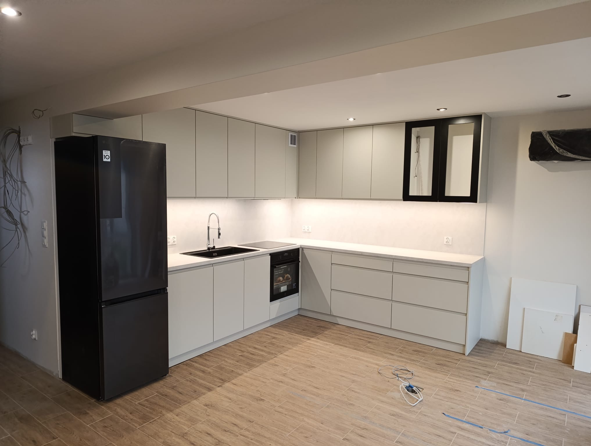 Jasna kuchnia z czarnym sprzętem AGD i drewnopodobną podłogą. Na ścianie widoczne przewody elektryczne. Minimalistyczny design i oświetlenie LED.