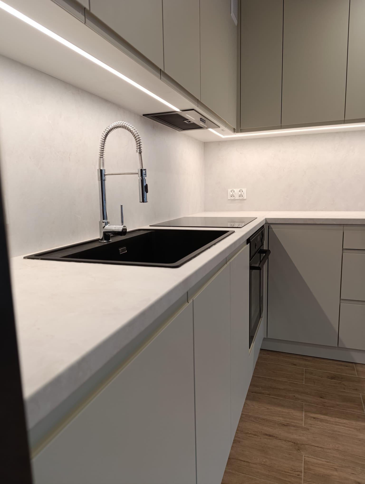 Nowoczesna kuchnia w odcieniach szarości z czarnym zlewem i baterią. Podświetlenie LED nad blatem. Minimalistyczny design i drewniana podłoga.