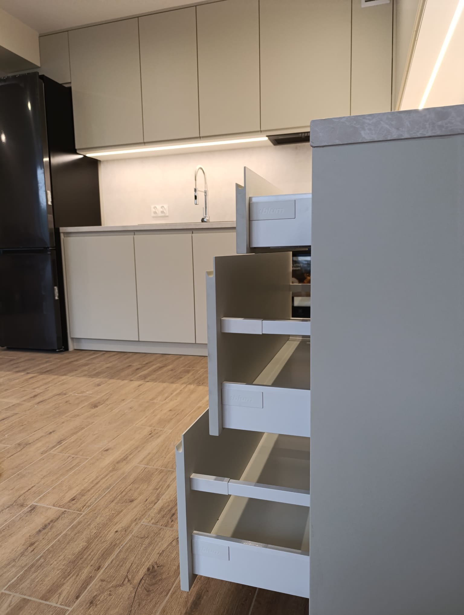 Nowoczesna kuchnia na wymiar z wysuwanymi szufladami Blum, widoczne w Czechowicach-Dziedzicach. Jasne fronty, drewnopodobna podłoga i czarna lodówka w tle.