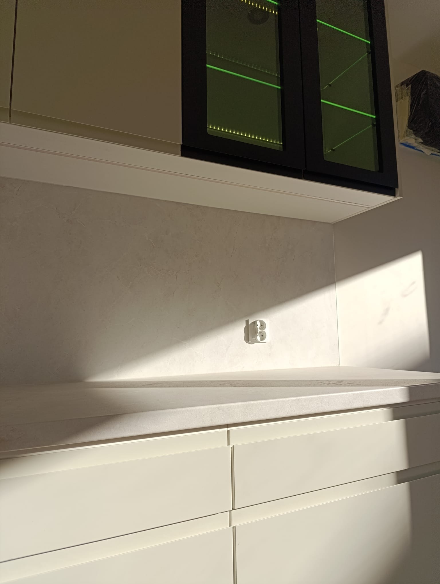 Jasna kuchnia: białe szafki z szufladami, marmurowy blat i górne szafki z podświetleniem LED. Na ścianie gniazdko w promieniach słońca.