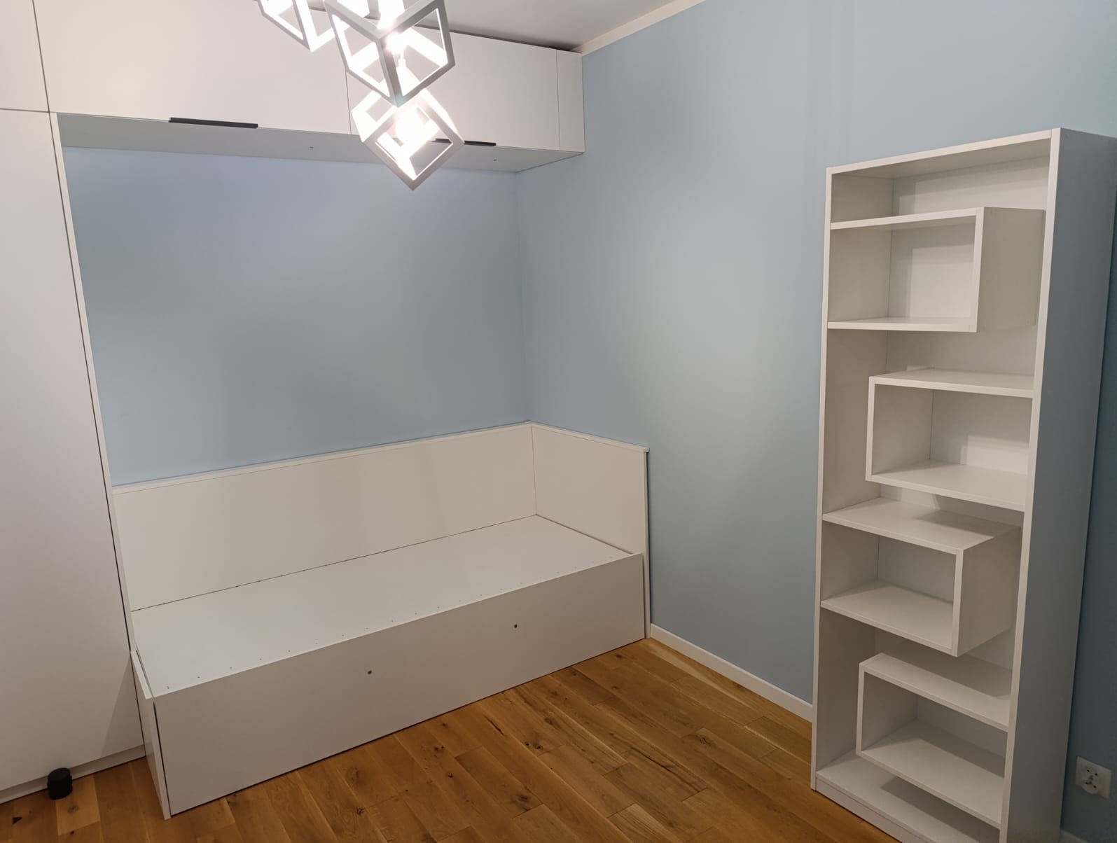 Nowoczesne wnętrze pokoju z zabudową meblową: biała szafa, narożna kanapa w zabudowie i regał o geometrycznym designie na drewnianej podłodze. Błękitna ściana.