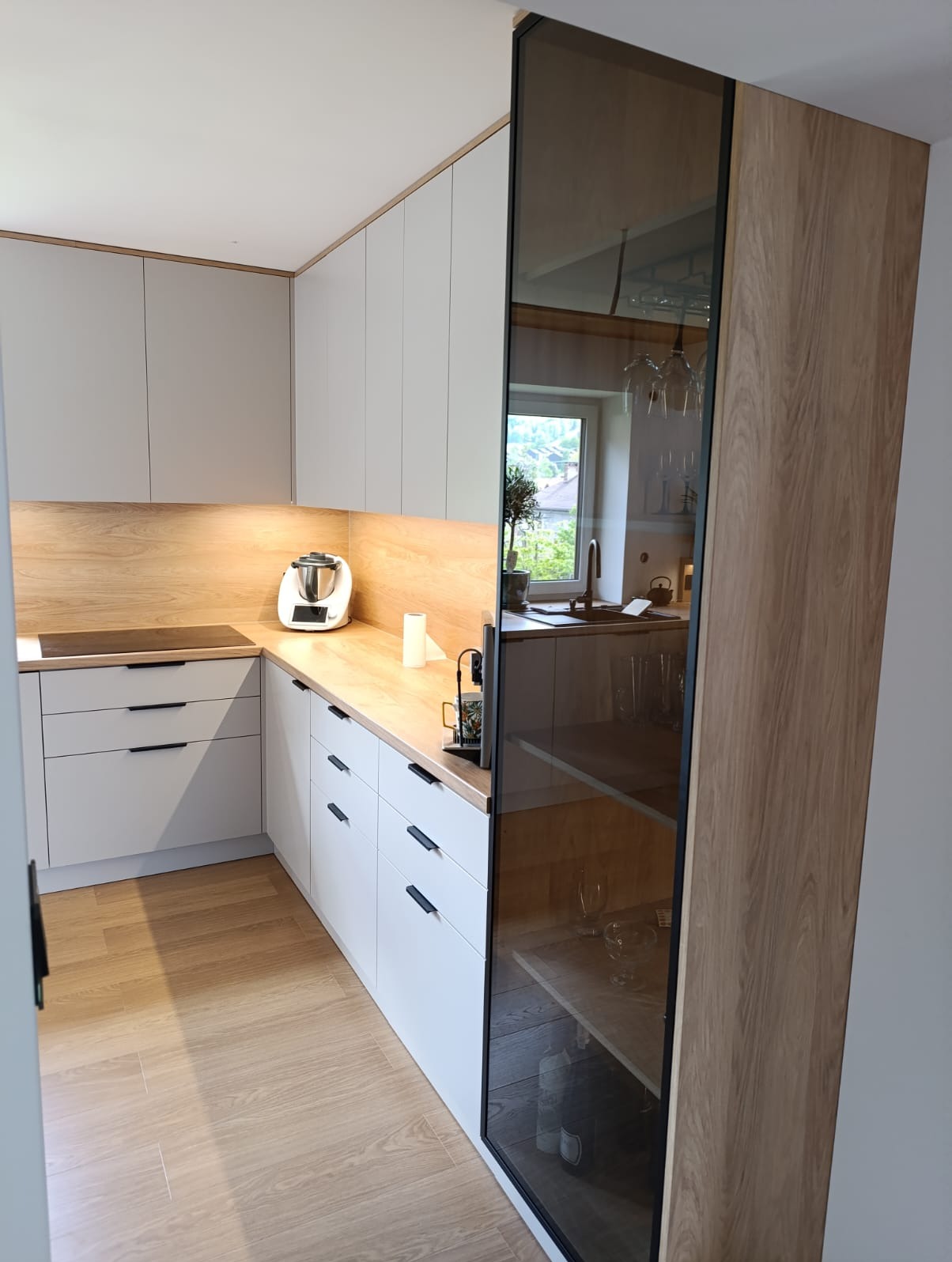 Nowoczesna kuchnia w odcieniach bieli i drewna, z minimalistycznymi uchwytami i szklaną witryną. Uwagę przyciąga drewniany blat i oświetlenie podszafkowe.