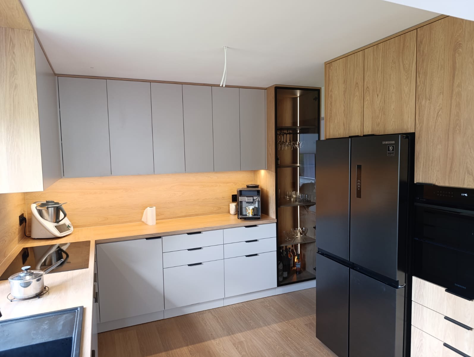 Nowoczesna kuchnia z szarymi frontami i drewnianymi akcentami, oświetleniem LED pod szafkami, czarną lodówką i barkiem z alkoholem.