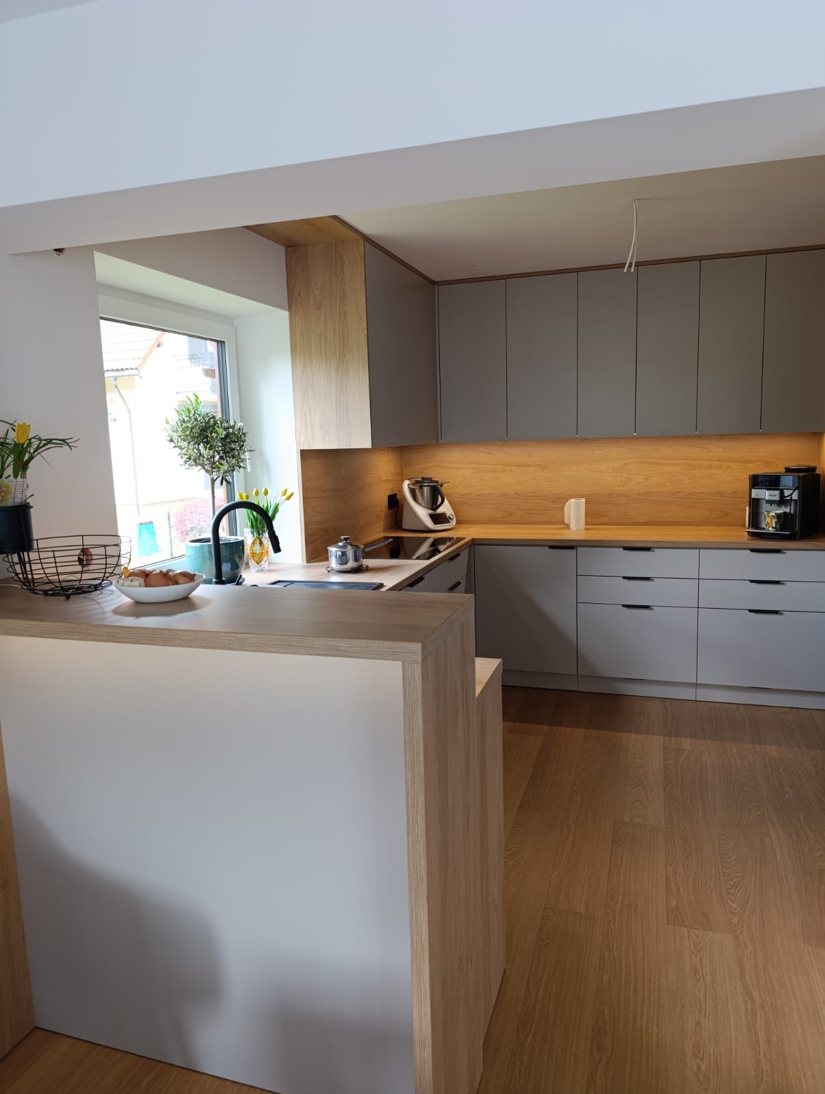 Nowoczesna kuchnia z szarymi szafkami, drewnianym blatem i wyspą. Na blacie kwiaty, owoce i akcesoria kuchenne. Oświetlenie LED pod szafkami.