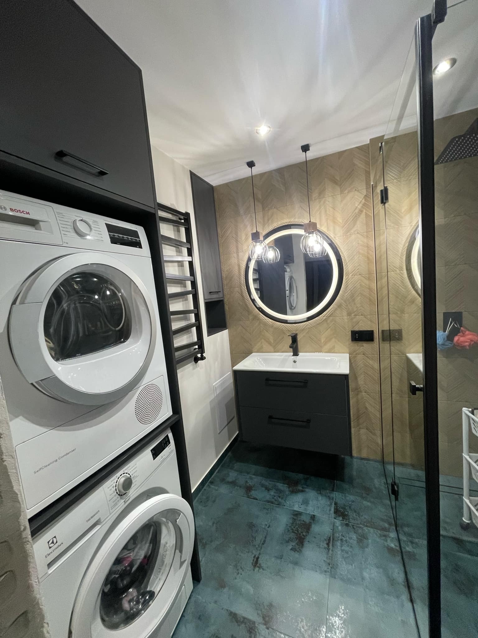 Nowoczesna łazienka z pralką i suszarką w zabudowie, okrągłym lustrem z podświetleniem i kabiną prysznicową. Ciemne meble, drewnopodobne płytki na ścianie.