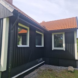 Gabriel Kłosiński - Elewacja domu z czarną, drewnianą fasadą, białymi ramami okiennymi i pomarańczową dachówką. Widoczny klimatyzator LG i kamienny element dekoracyjny w ogrodzie.