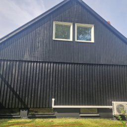 Gabriel Kłosiński - Ciemna, drewniana elewacja domu z dwoma oknami na poddaszu, widok z zewnątrz. Na dole widoczne jednostki klimatyzacyjne i fragment trawnika.