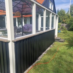 Gabriel Kłosiński - Ciemna, odnowiona elewacja oranżerii z białymi ramami okiennymi, widok z zewnątrz na zadbany trawnik i elementy wyposażenia wnętrza.