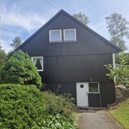 Gabriel Kłosiński - Dom z czarną, drewnianą elewacją i białymi oknami, otoczony zielenią. Widoczny fragment ścieżki i flaga na maszcie. Skandynawski styl.