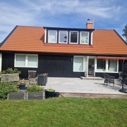 Gabriel Kłosiński - Czarny dom z pomarańczową dachówką, drewnianym tarasem i elementami dekoracyjnymi na zewnątrz. Widok na ogród z roślinami w donicach i meblami ogrodowymi.