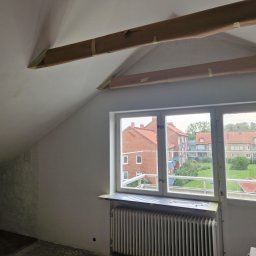 Gabriel Kłosiński - Poddasze w trakcie przygotowań do malowania: zabezpieczone taśmą malarską belki stropowe i parapet okienny. Widok na budynki przez okno.