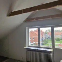 Poddasze w trakcie przygotowań do malowania: zabezpieczone taśmą malarską belki stropowe i parapet okienny. Widok na budynki przez okno.