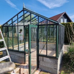 Gabriel Kłosiński - Szklarnia ogrodowa z otwartymi drzwiami, zbudowana na fundamencie z szarego betonu komórkowego. Drabina oparta o bok. Błękitne niebo w tle.