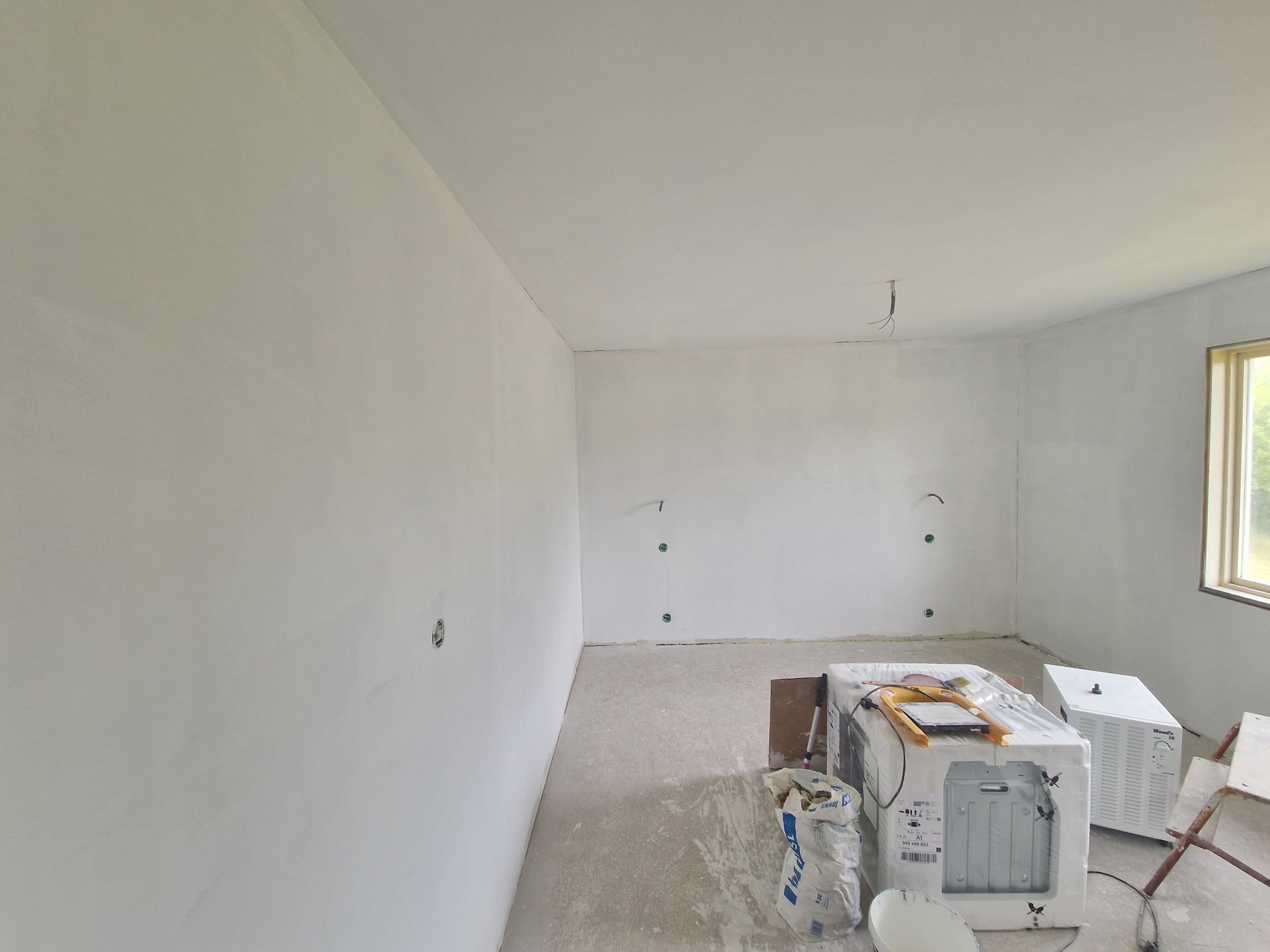 Wnętrze po szpachlowaniu, ściany i sufit pokryte gładzią, widoczne puszki elektryczne. Na podłodze worki z materiałami i sprzęt budowlany. Okno z widokiem na zieleń.