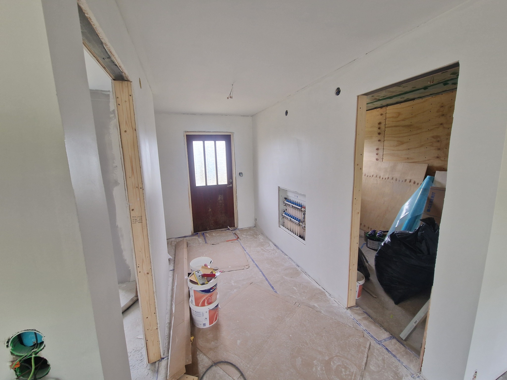 Wnętrze w trakcie remontu: świeżo wyszpachlowane ściany, widoczne puszki z farbą, niezabezpieczone futryny drzwi i rozdzielacz centralnego ogrzewania.