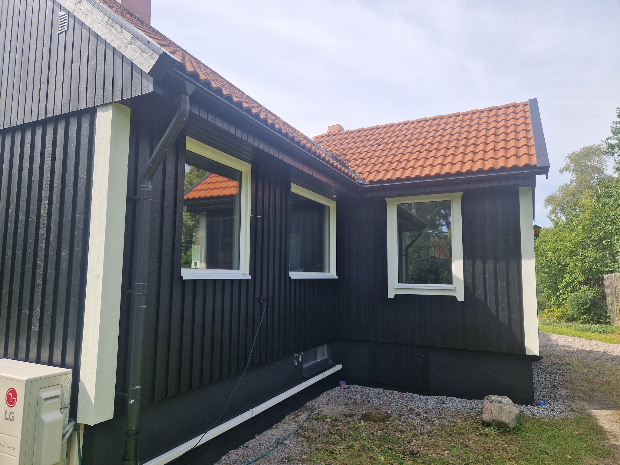 Elewacja domu z czarną, drewnianą fasadą, białymi ramami okiennymi i pomarańczową dachówką. Widoczny klimatyzator LG i kamienny element dekoracyjny w ogrodzie.