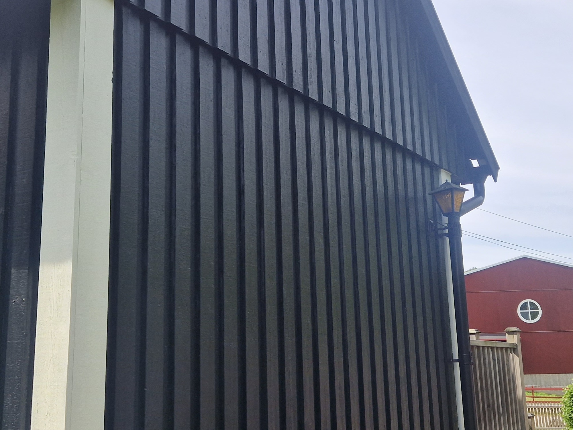 Ciemna, drewniana elewacja budynku z pionowymi deskami, oświetlona lampą. Białe wykończenia narożników. W tle czerwony budynek z okrągłym oknem.