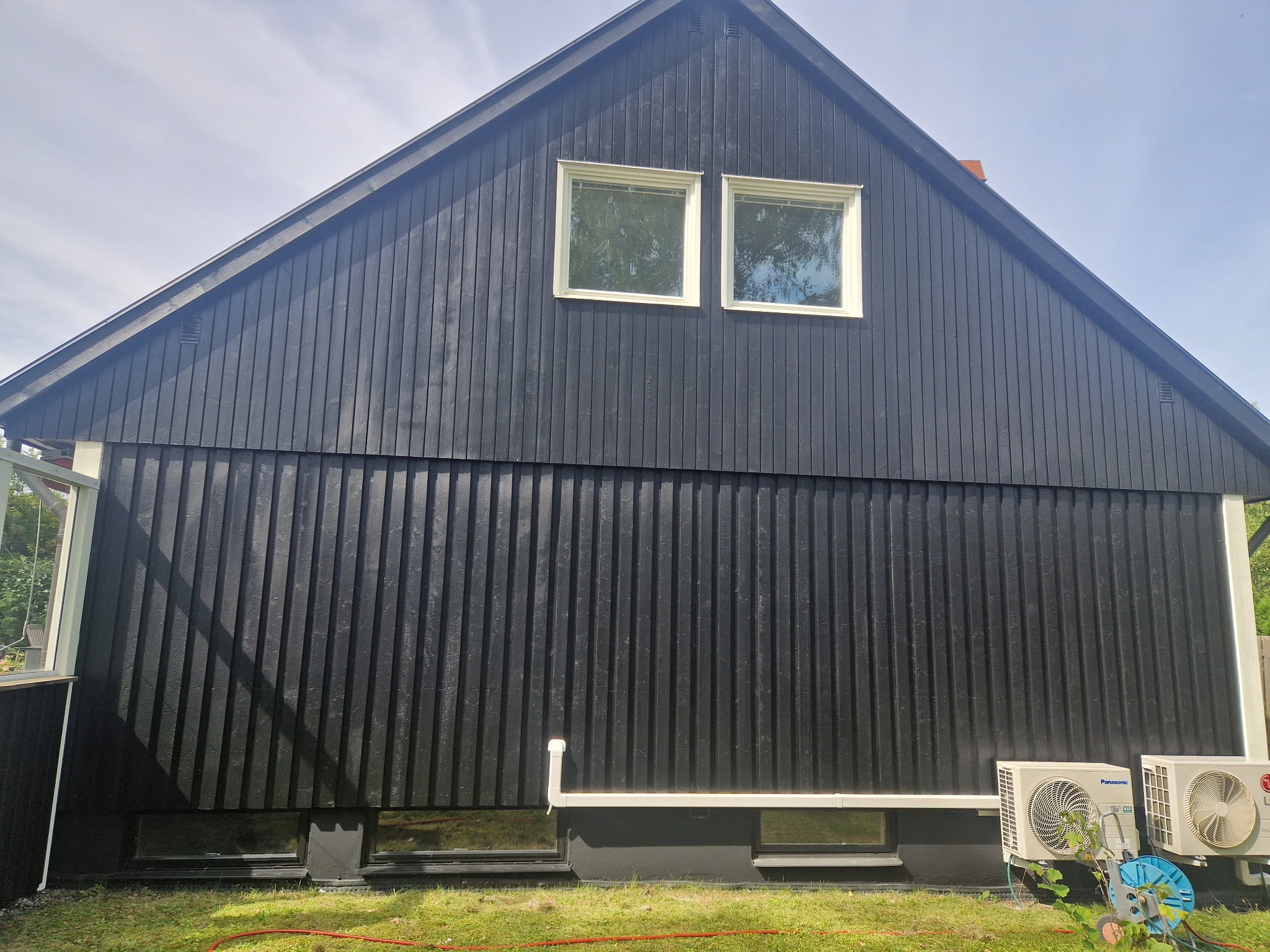 Ciemna, drewniana elewacja domu z dwoma oknami na poddaszu, widok z zewnątrz. Na dole widoczne jednostki klimatyzacyjne i fragment trawnika.
