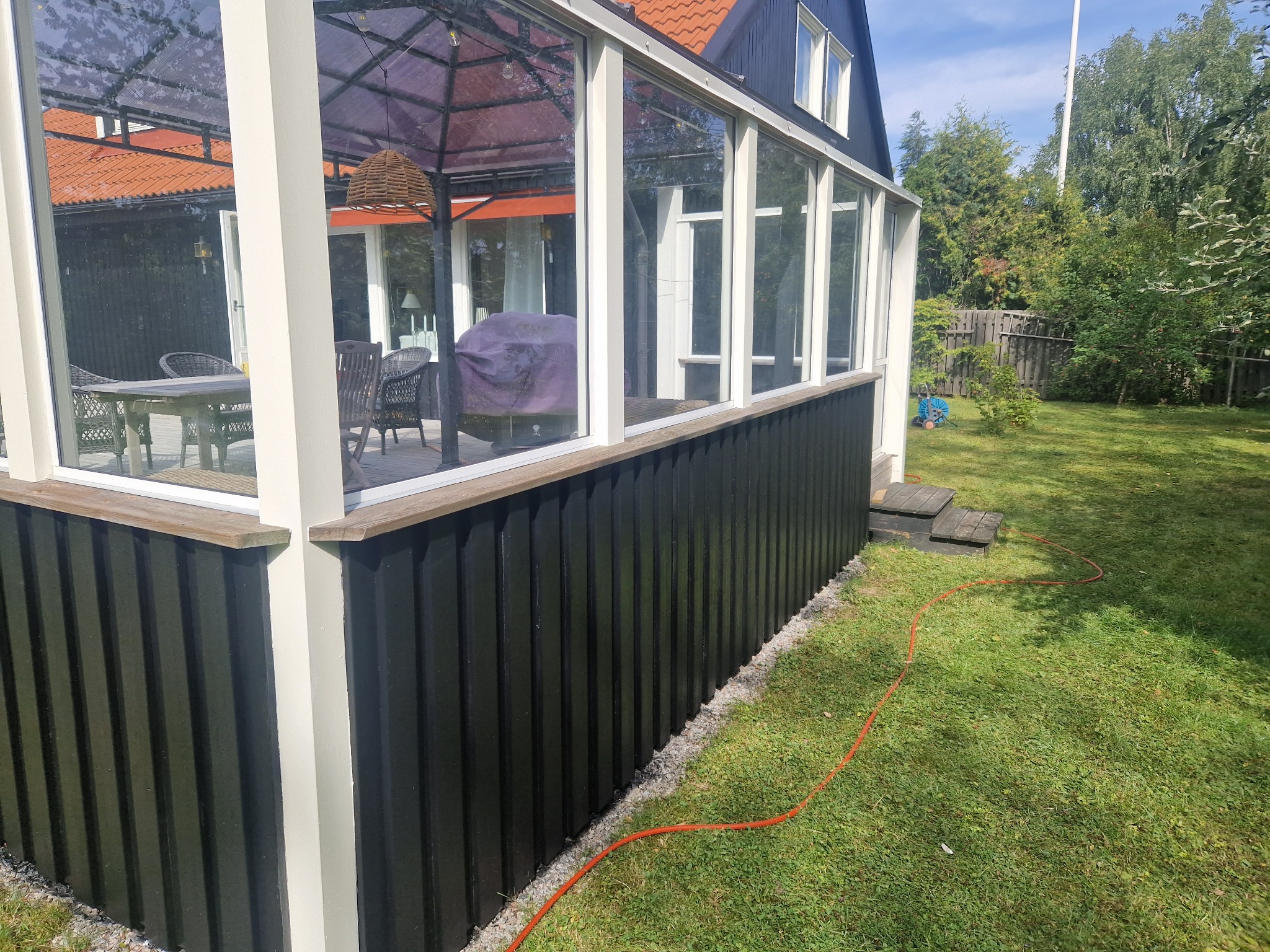 Ciemna, odnowiona elewacja oranżerii z białymi ramami okiennymi, widok z zewnątrz na zadbany trawnik i elementy wyposażenia wnętrza.