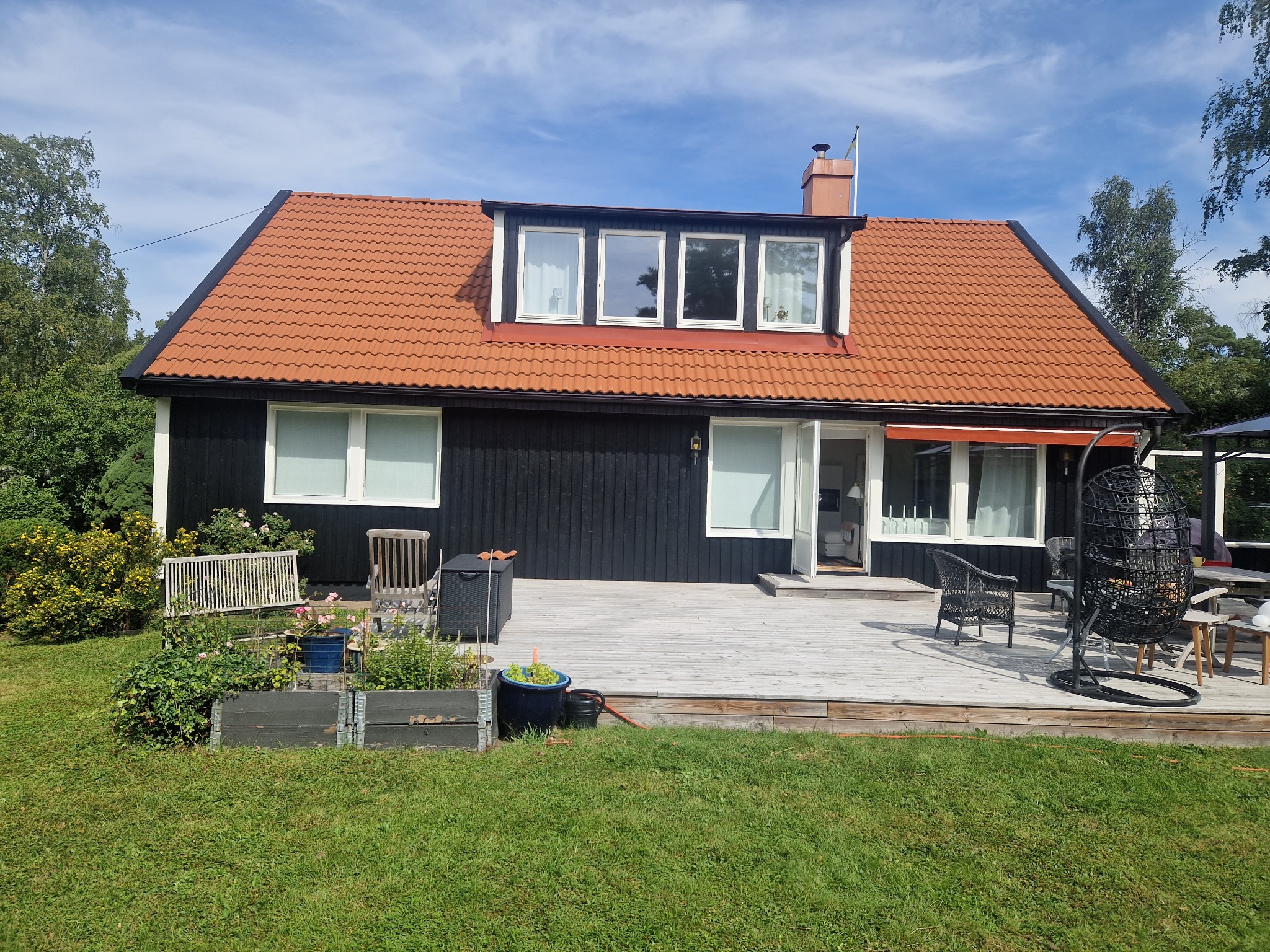 Czarny dom z pomarańczową dachówką, drewnianym tarasem i elementami dekoracyjnymi na zewnątrz. Widok na ogród z roślinami w donicach i meblami ogrodowymi.