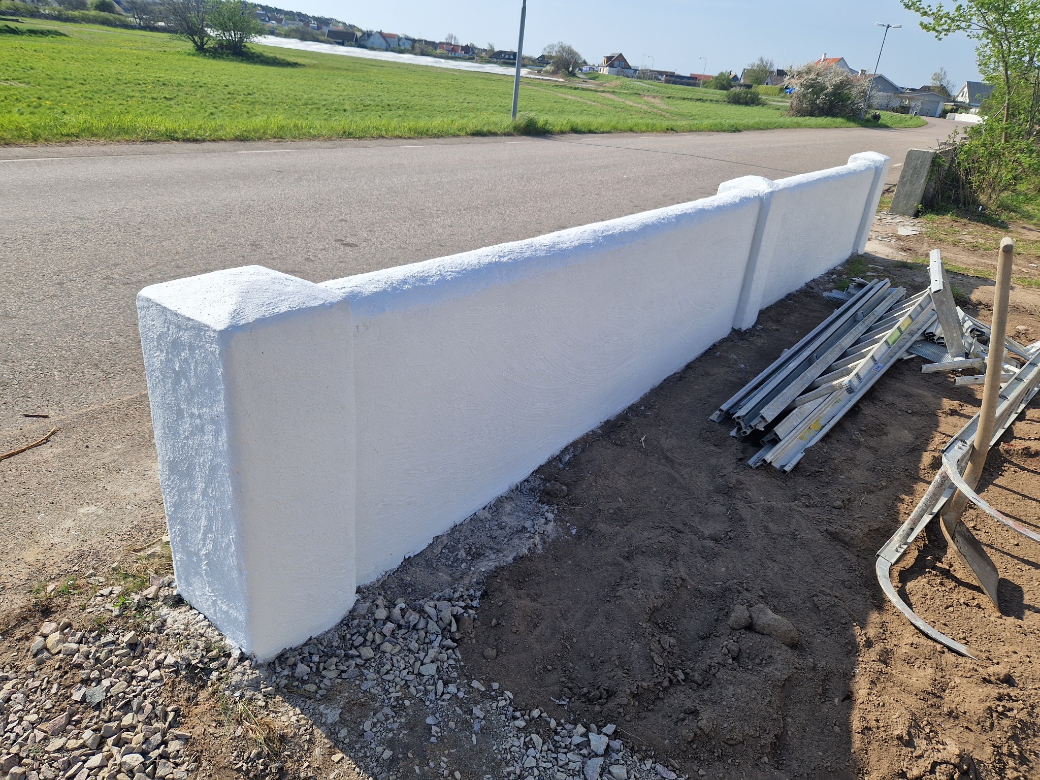 Świeżo otynkowany, biały murek oddzielający posesję od drogi, z widocznymi narzędziami. Tło: zielony krajobraz z zabudowaniami. Jasne, słoneczne światło.