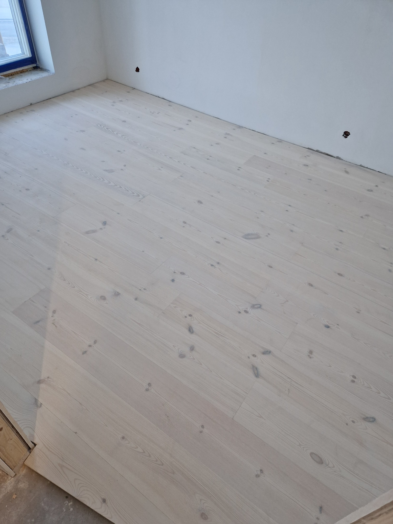 Jasna podłoga z paneli ułożona w pokoju z białymi ścianami i oknem. Widoczne otwory w ścianach na wysokości podłogi. Fragment progu w lewym dolnym rogu.