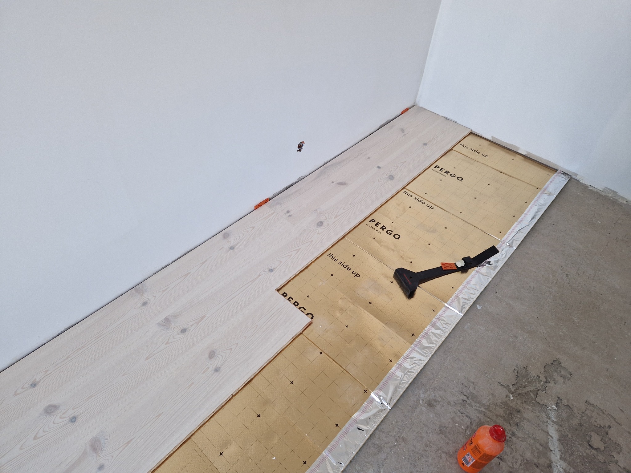 Białe panele podłogowe Pergo w trakcie montażu, widoczna podkładka izolacyjna, narzędzie do docinania i butelka z klejem. Ściany białe, surowy beton na podłodze.