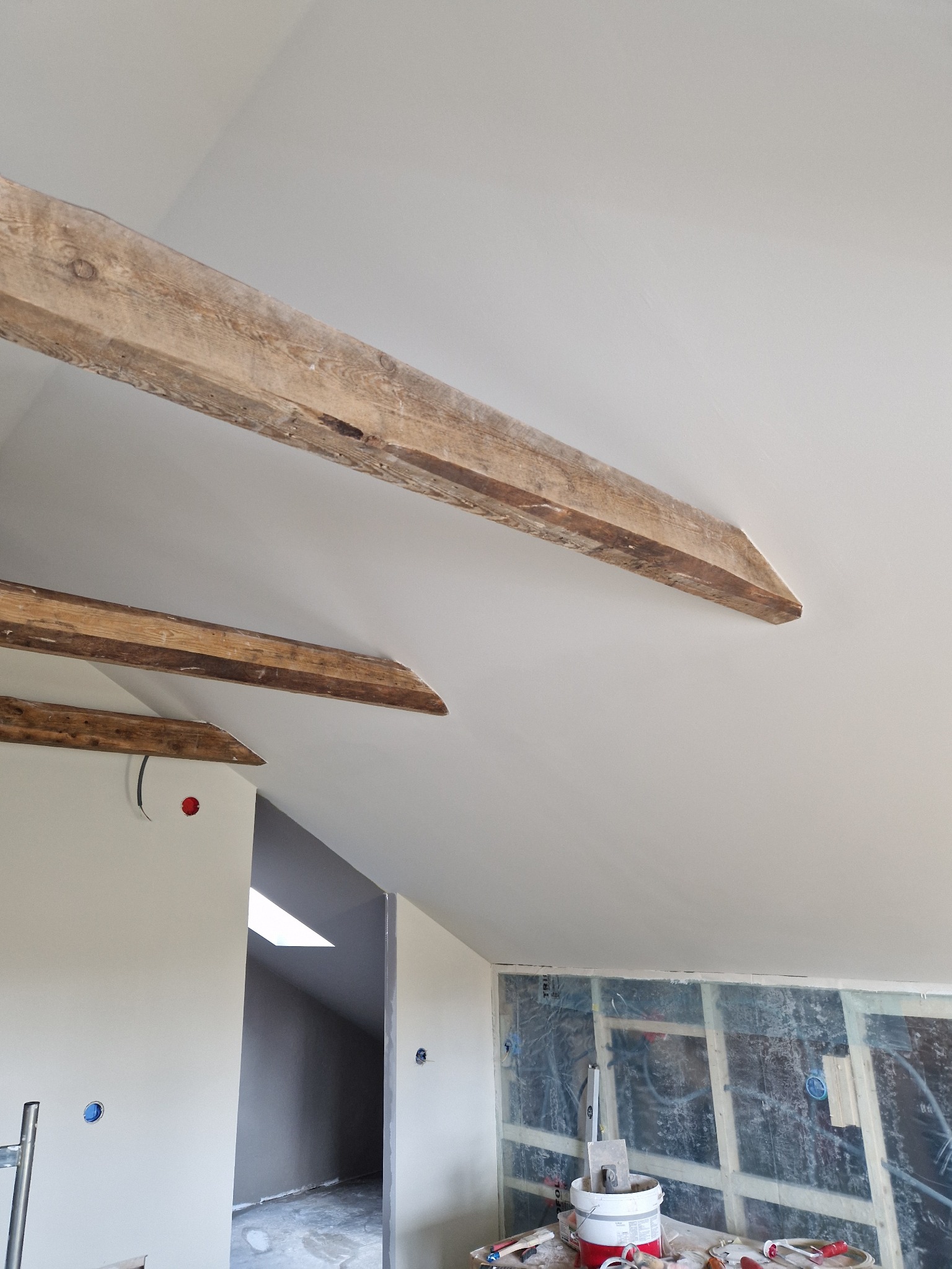 Biały sufit z drewnianymi belkami w remontowanym wnętrzu. Widoczne narzędzia i materiały budowlane. Ściany przygotowane do dalszych prac wykończeniowych.