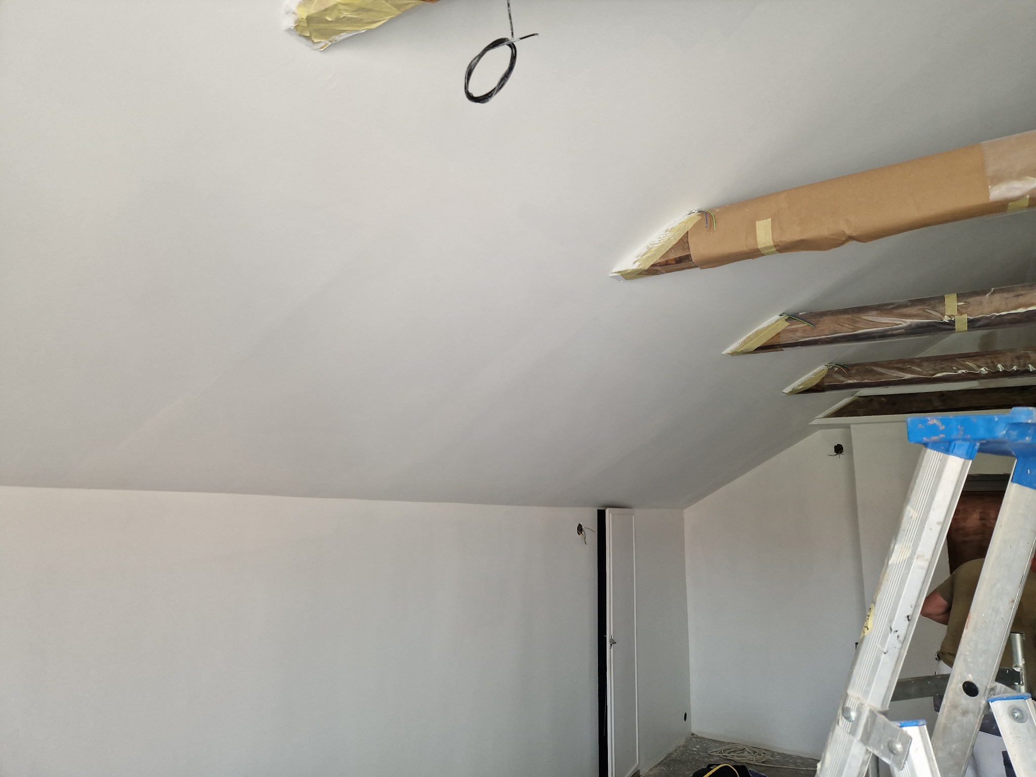 Białe ściany i sufit po malowaniu w poddaszu ze skosami. Widoczne zabezpieczone elementy drewniane i drabina malarska.