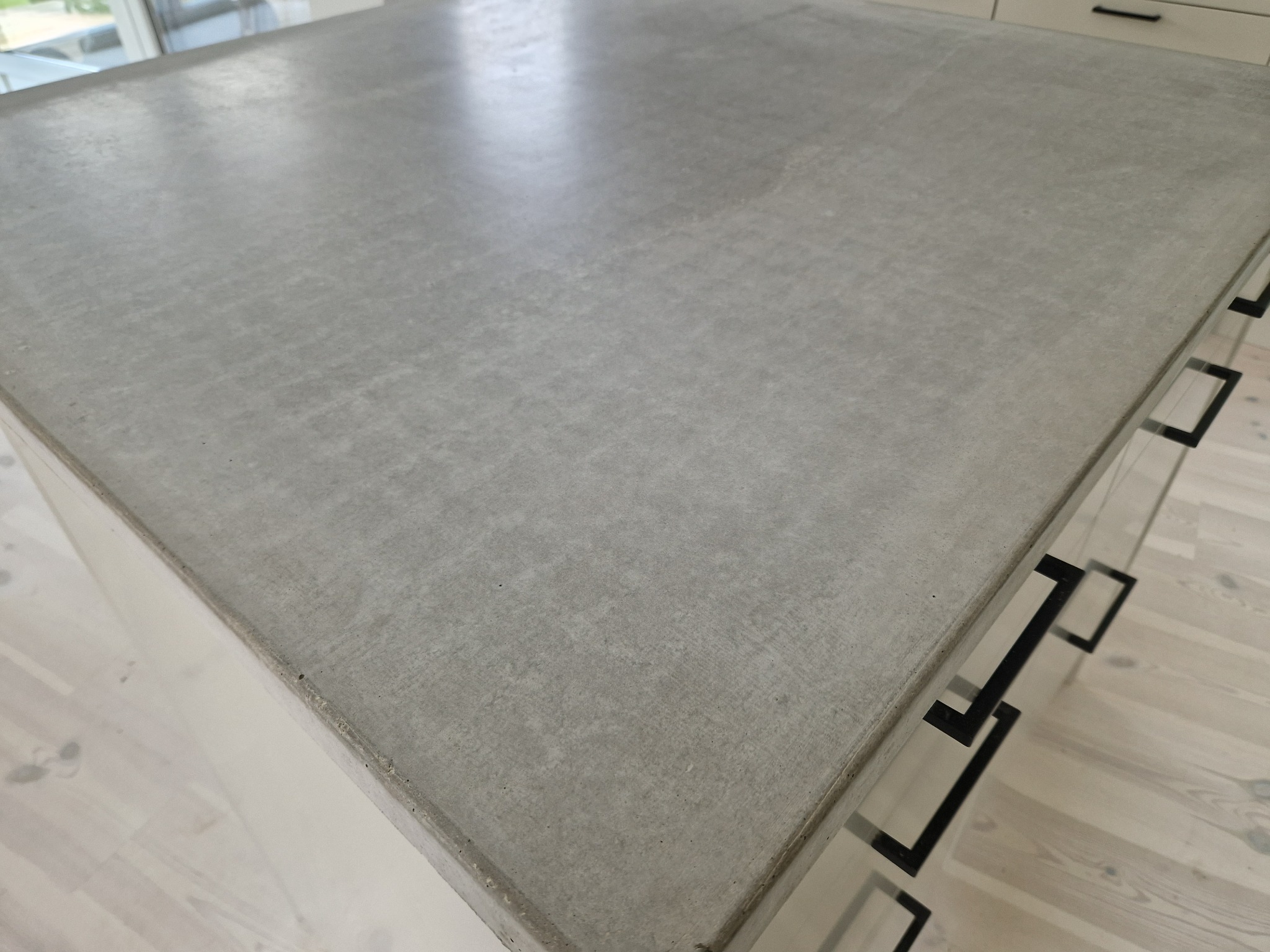 Gładki, szary blat betonowy wyspy kuchennej z czarnymi uchwytami szafek, widoczny fragment drewnianej podłogi, nowoczesny design.