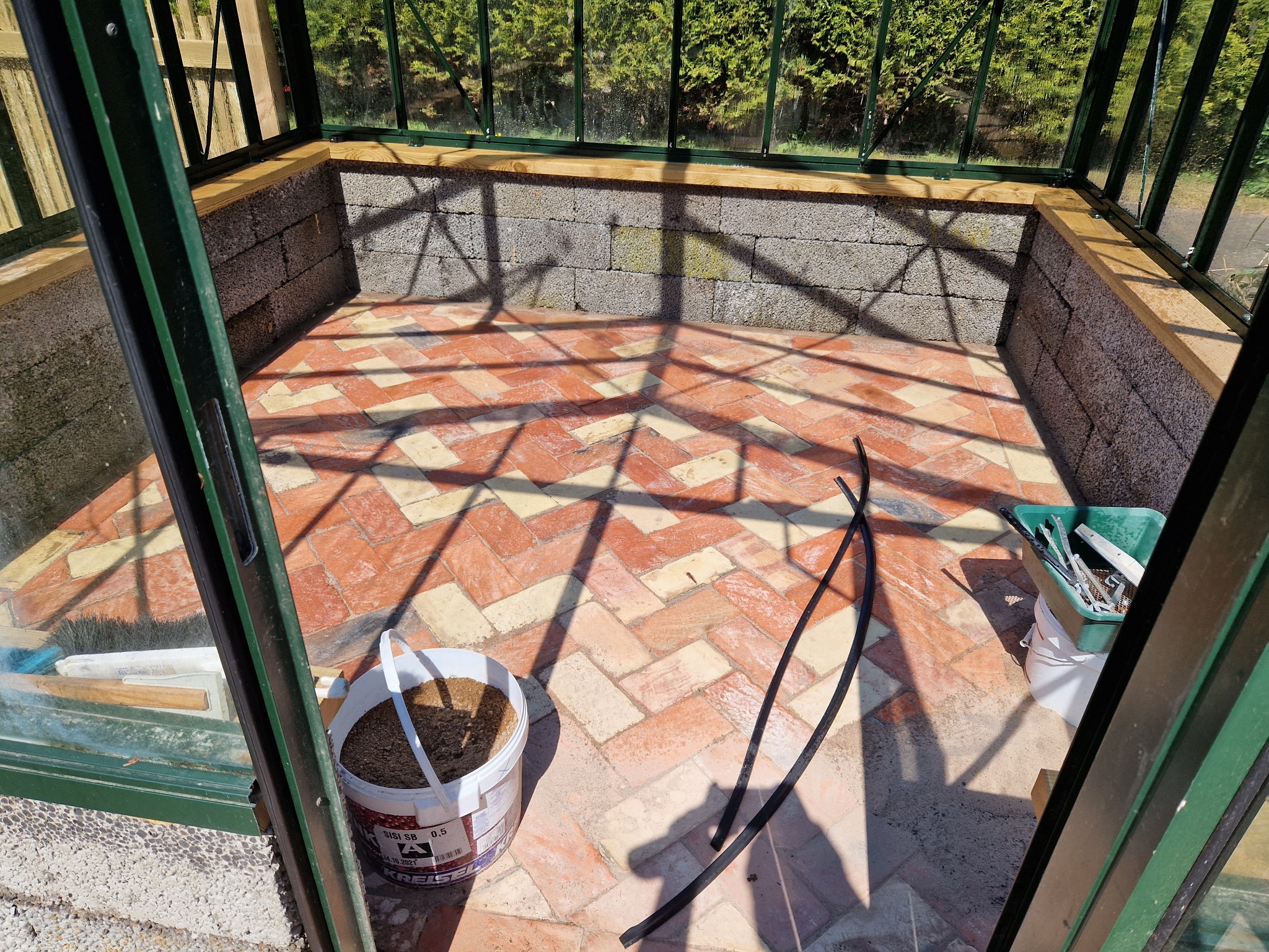 Wnętrze szklarni z podłogą z cegły ułożonej w jodełkę, ściany z szarego betonu komórkowego, widoczne narzędzia i wiadro z ziemią, oświetlone słonecznym światłem.