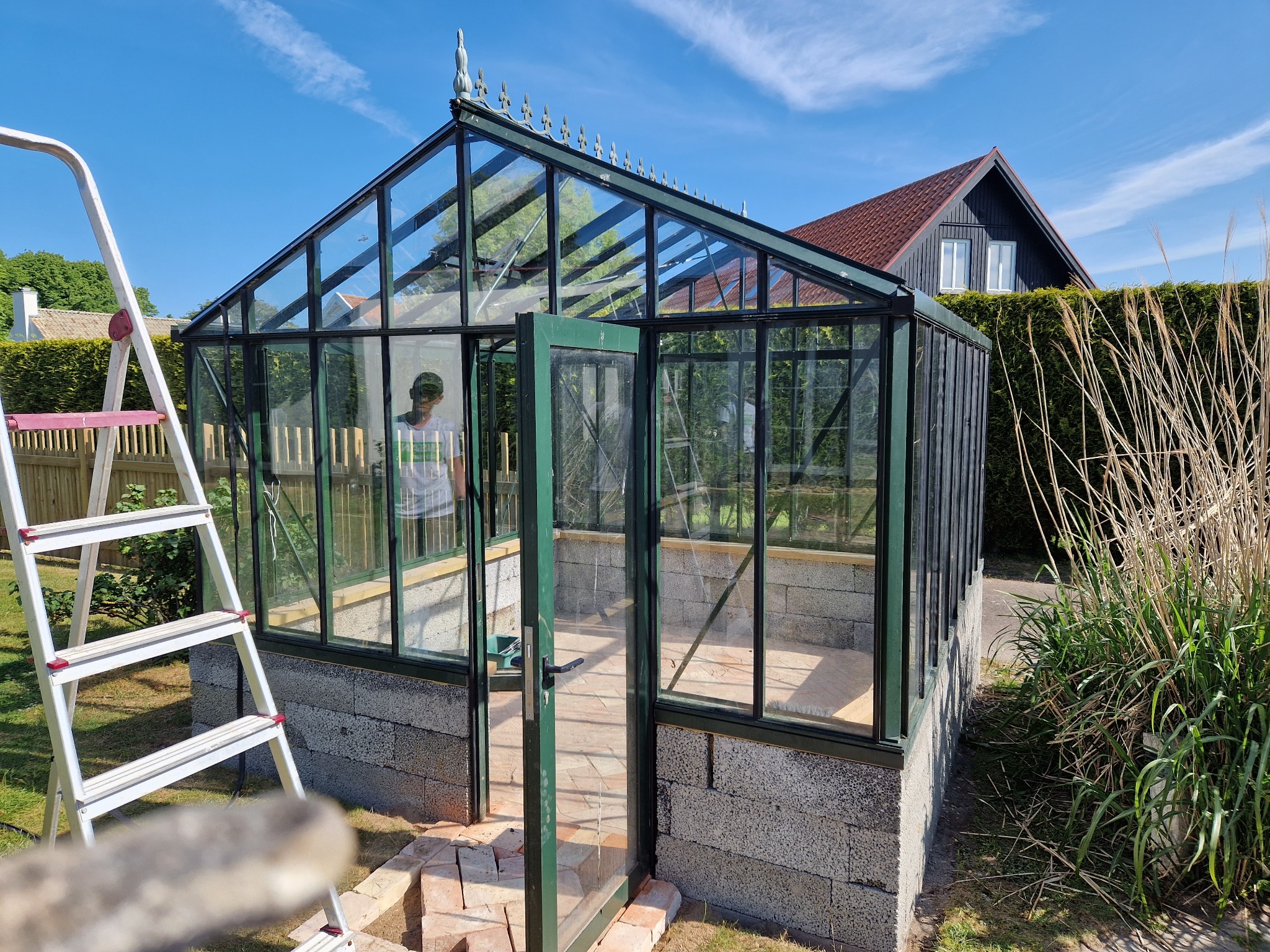 Szklarnia ogrodowa z otwartymi drzwiami, zbudowana na fundamencie z szarego betonu komórkowego. Drabina oparta o bok. Błękitne niebo w tle.