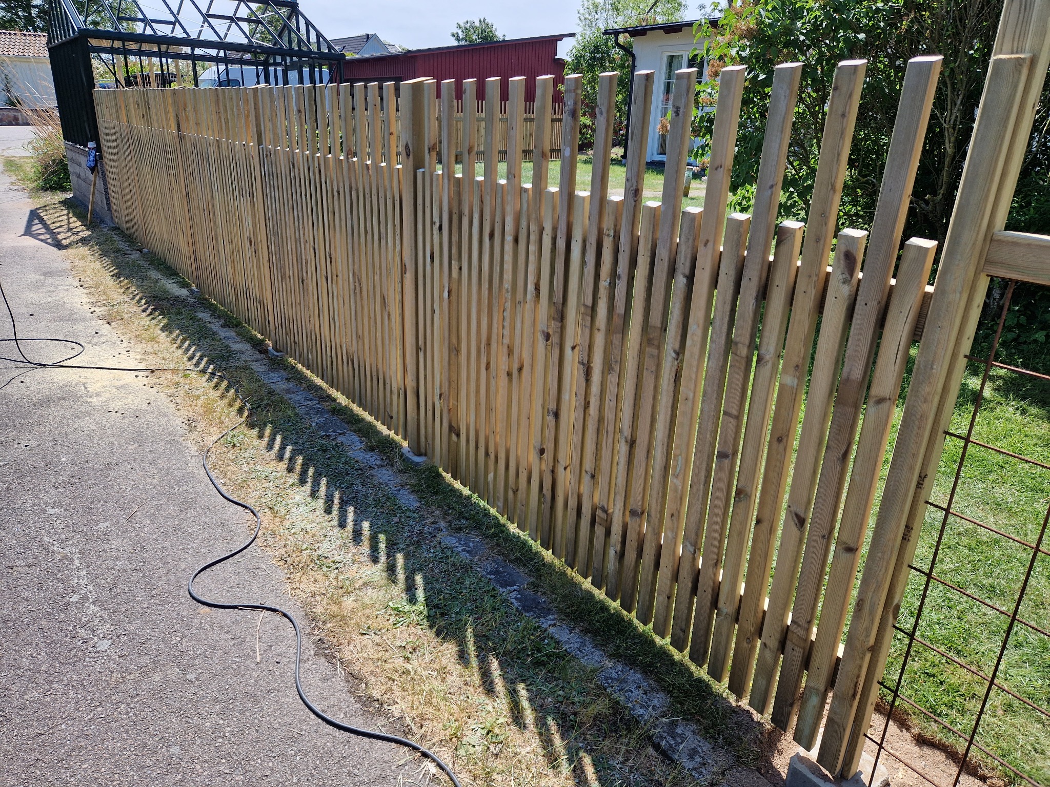 Nowoczesne ogrodzenie z pionowych listew drewnianych, z przerwami, na tle zieleni ogrodu i budynku z czerwonym dachem, z widocznym kablem narzędzia.