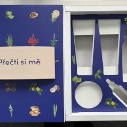 Niebieskie opakowanie z motywami roślinnymi, zawierające wycięcia na kosmetyki, z tekstem 'Přečti si mě' na beżowej wkładce.