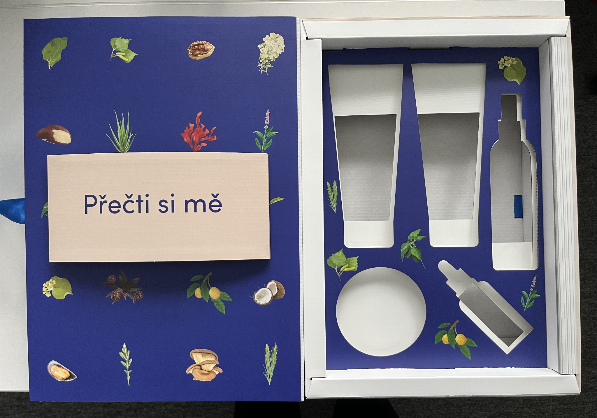 Niebieskie opakowanie z motywami roślinnymi, zawierające wycięcia na kosmetyki, z tekstem 'Přečti si mě' na beżowej wkładce.