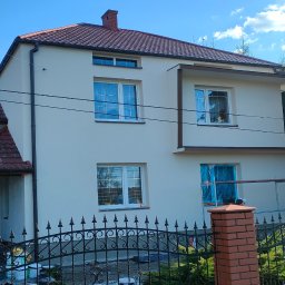 Konrad Sowiński usługi remontowo budowlane - Dom po remoncie elewacji w Górze, widoczny przez ozdobne ogrodzenie. Świeżo pomalowana fasada w jasnym kolorze, z ciemnobrązowymi detalami. Częściowo widoczne rusztowania.