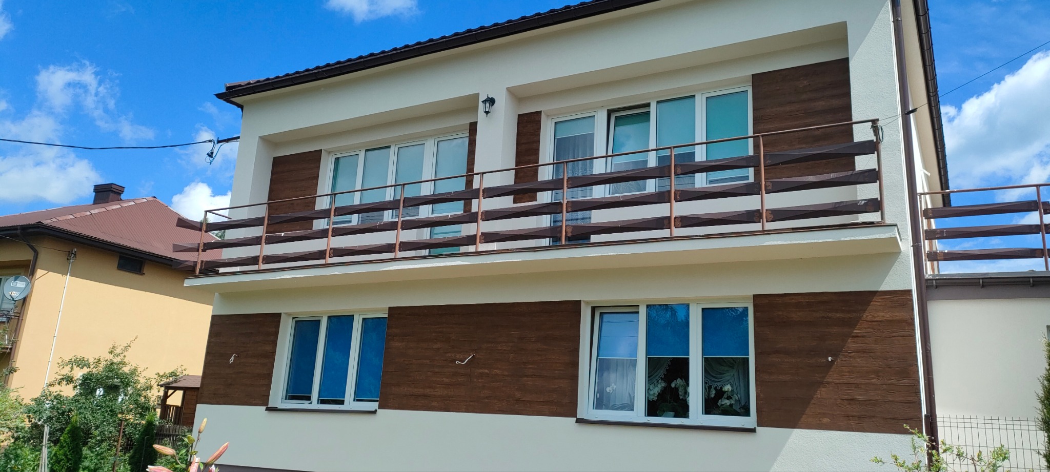 Dwukondygnacyjny dom po renowacji elewacji, z drewnopodobną okładziną i balkonem z ozdobną balustradą, na tle błękitnego nieba.