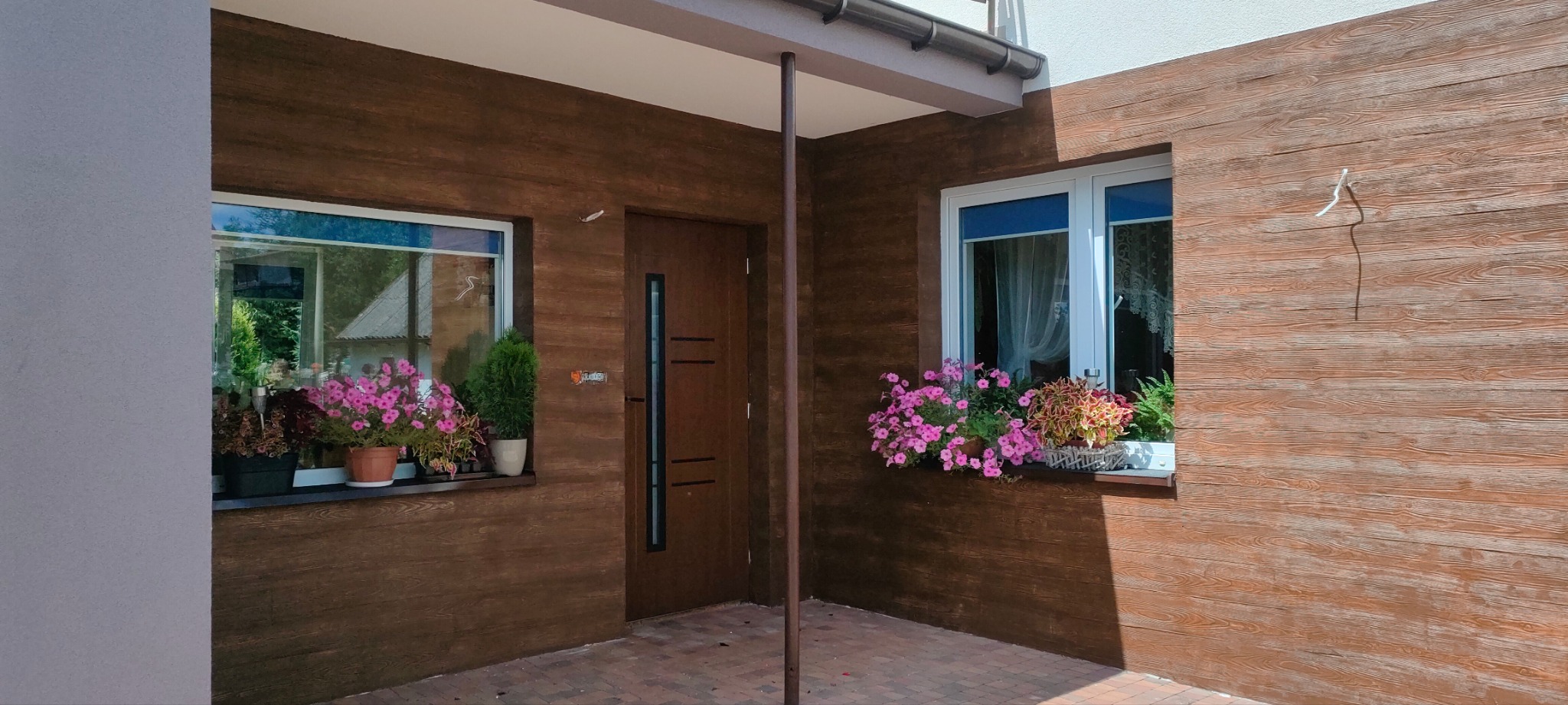 Elewacja domu z fakturą drewna, okna z kwiatami w doniczkach, brązowe drzwi z przeszkleniem. Eleganckie wykończenie zewnętrzne budynku.