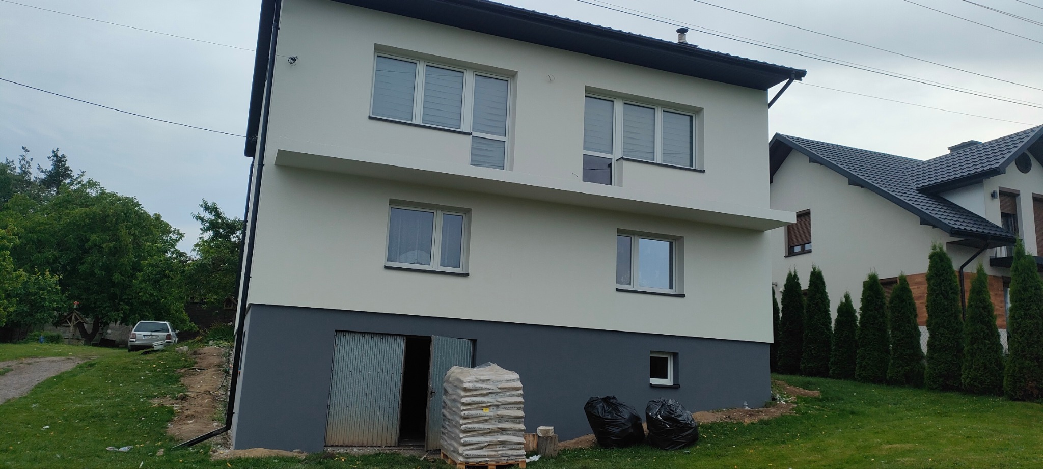 Dwukondygnacyjny dom z jasną elewacją i ciemnoszarą lamperią. Worki z materiałem budowlanym przed otwartymi drzwiami do piwnicy.