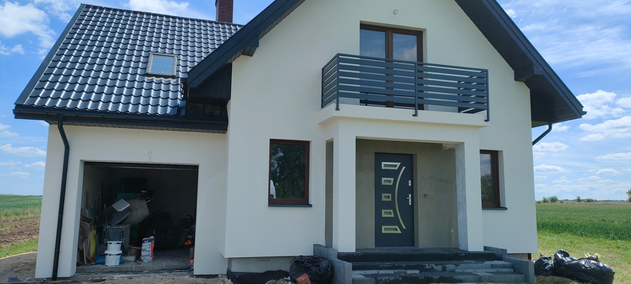 Biały dom z szarą stolarką i balkonem, widok z zewnątrz na otwarty garaż z narzędziami, fragment zielonego pola w tle, nie do końca wykończona elewacja.